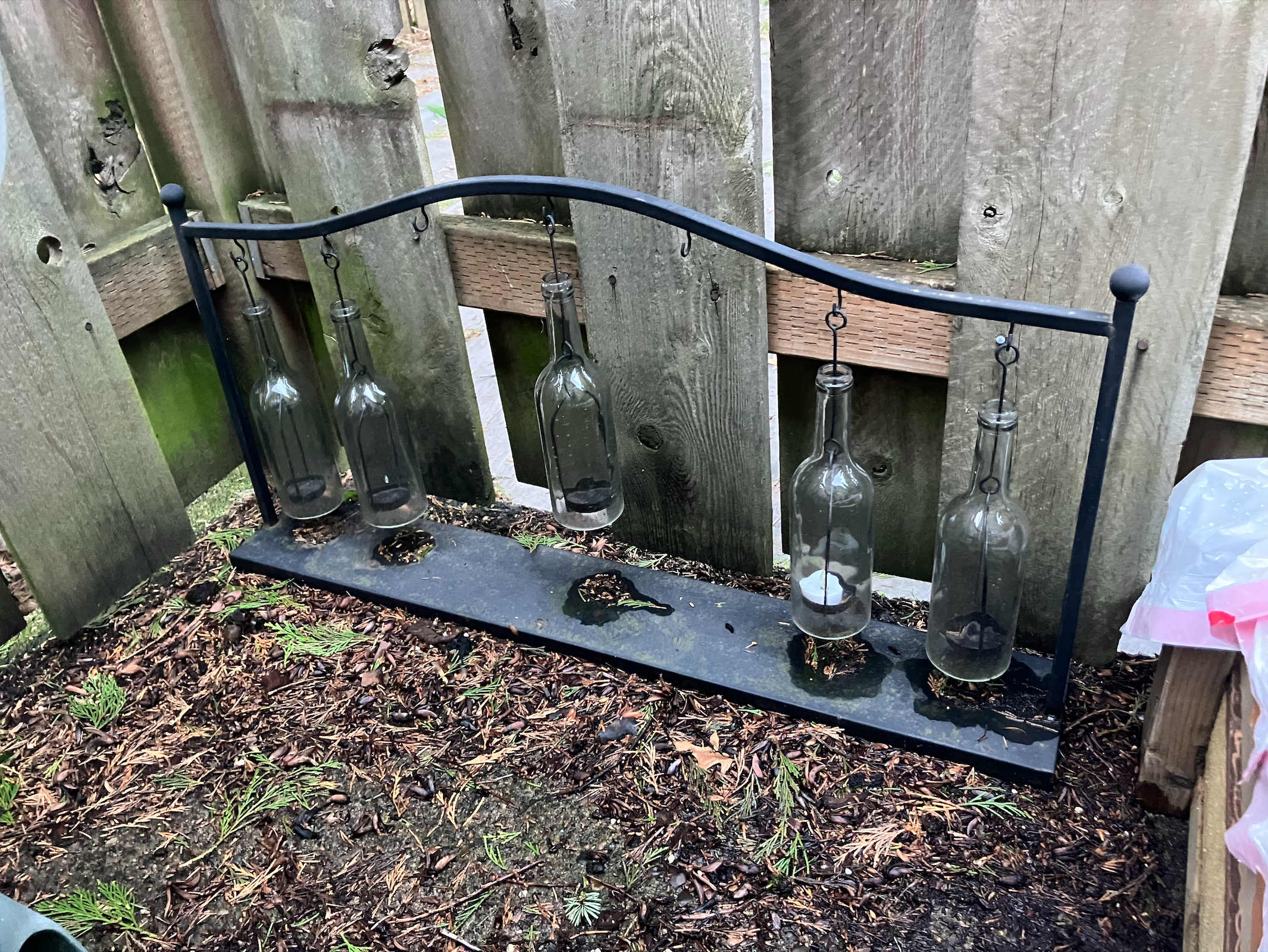 A cast iron candle holder with hanging glass bottles in front of a corner fence in an outdoor paved patio, with moss, pine needles and leaves on the ground around it.